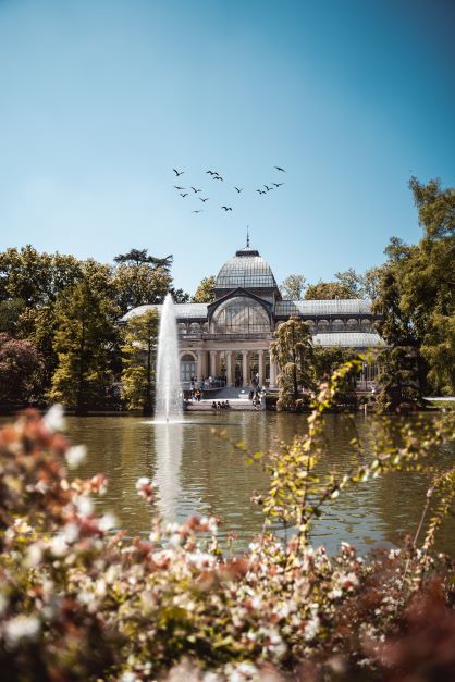 A beautiful photo of the glass palace in Retiro Park in Madrird surrounded by flowers and a beautiful fountain.. one of the best places to visit in Madrid