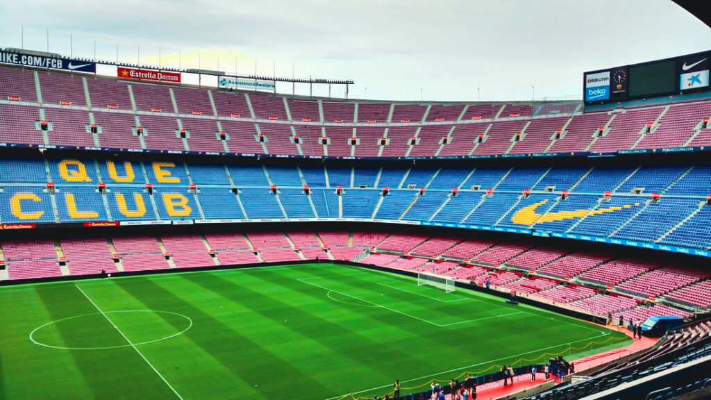 A wide angle photo of Camp Nou stadium with blue, pink and brown seats and a green field