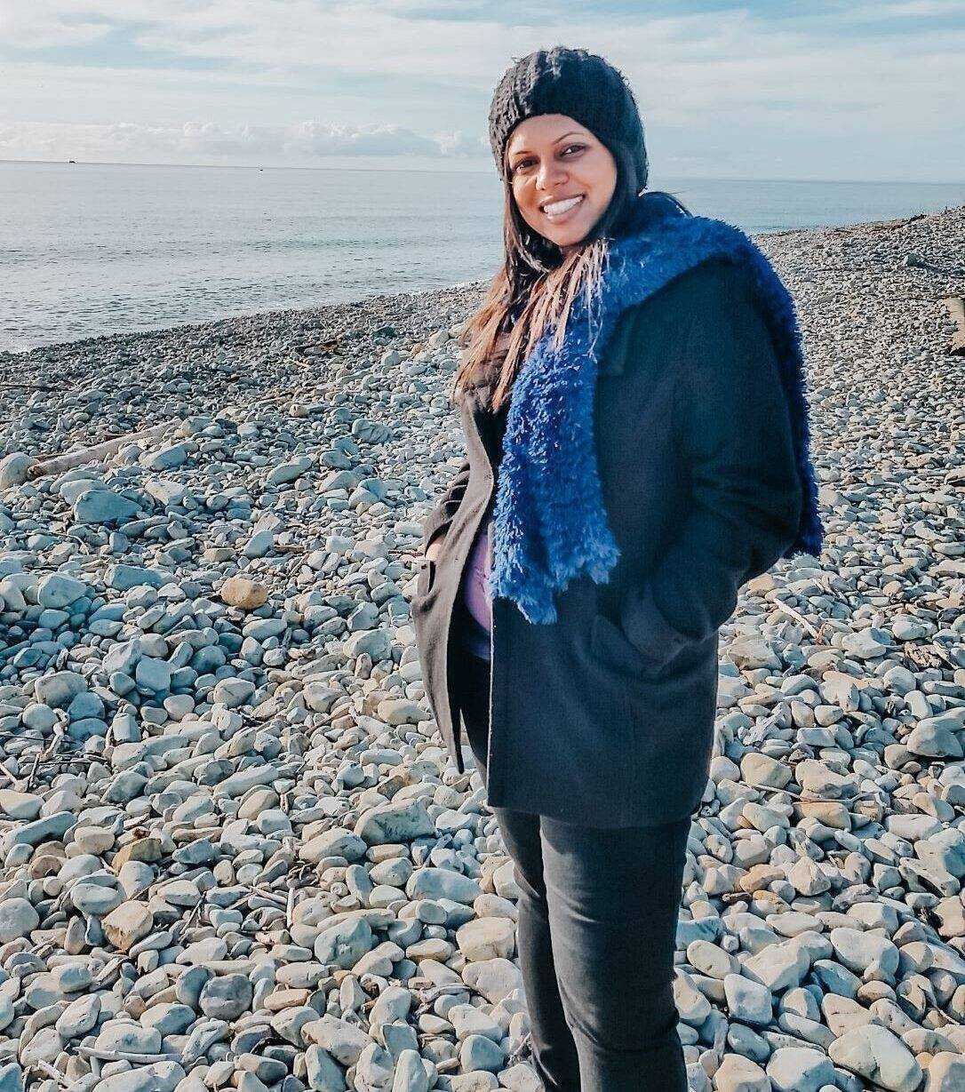 A picture of a woman in a winter coat, scarf and beanie standing by the water