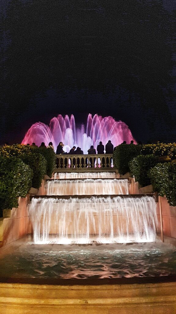 A photo showing a small waterfall and fountain lit up in pink and purple colours. An audience watching the fountain show