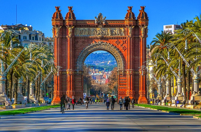 A brown concrete archway standing along a wide walkway with green palm trees on either side