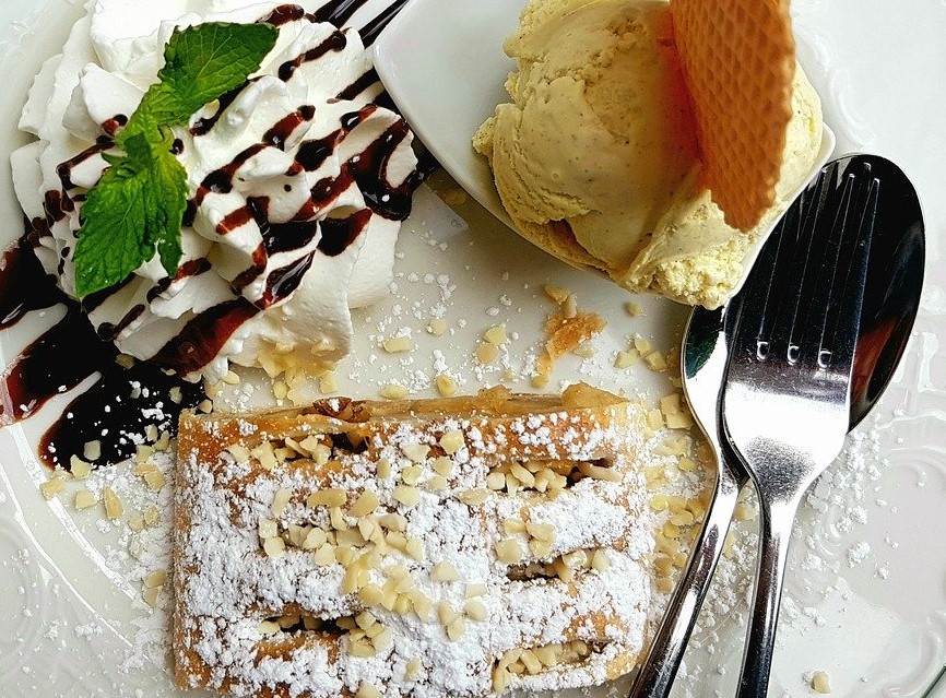 A plate of delicious looking apple strudel, dusted in icing sugar,topped with chopped nuts served with a scoop of ice-cream and fresh cream drizzled with chocolate sauce and garnished with mint. Would you say this is one of the best foods to eat in Vienna.