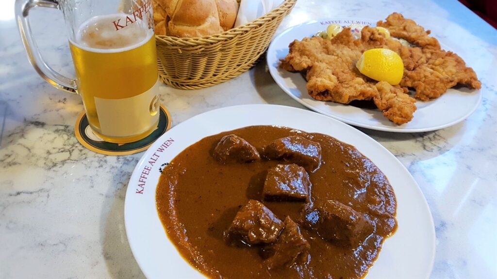 A plate of Beef Goulash in a thick sauce,  with a glass of cold beer, schnitzel and bread rolls in the background.