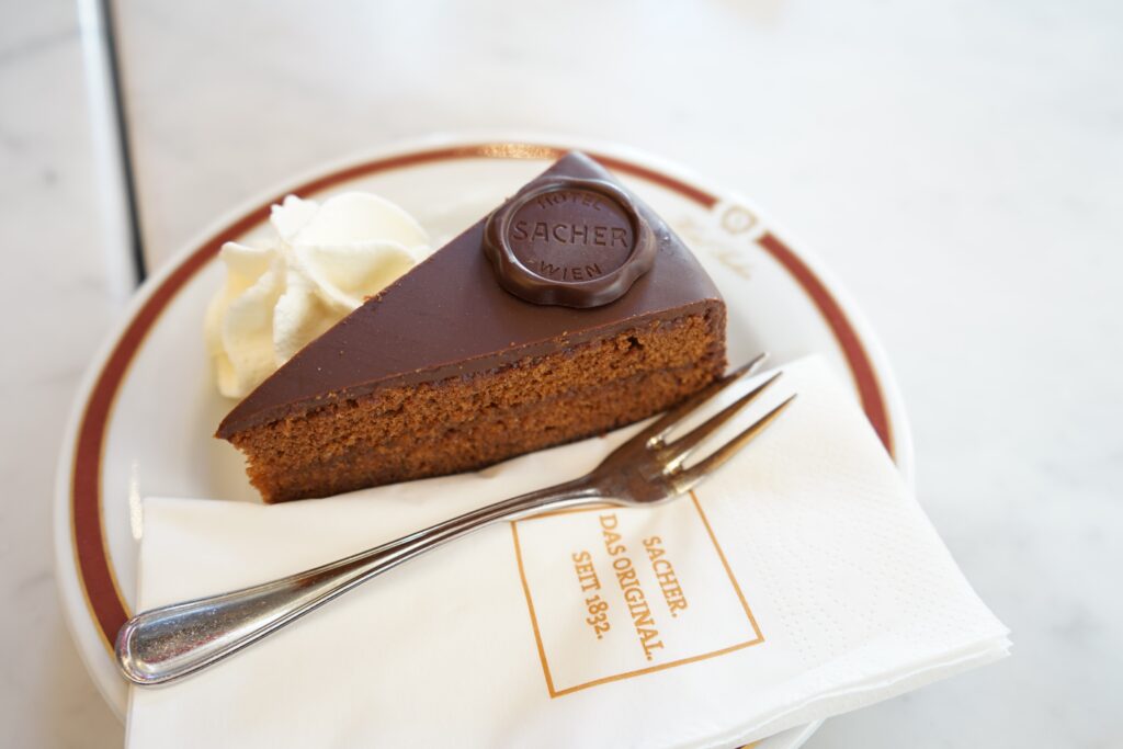 A triangular shaped slice of Sachertorte cake placed on a plate with a fork, serviette and whipped cream. This is one of the most traditional food in Austria you will find.