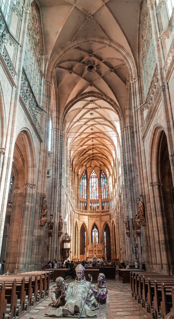 The interior of St Vitus Cathedral with high ceilings and intricate work on them. A statue sits right at the entrance of the walkway with pews on either side looking to the alter.