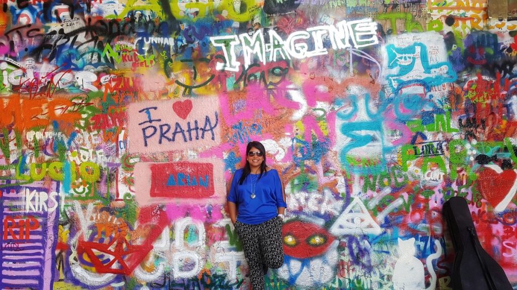 A picture of a colorful wall popularly known as the John Lennon Wall filled with graffiti and pieces of lyrics from the Beatles song. Writing on the wall includes "i love Praha" and a girl standing by the wall with a guitar resting against the wall. You cannot miss this top attractions to visit in Prague.