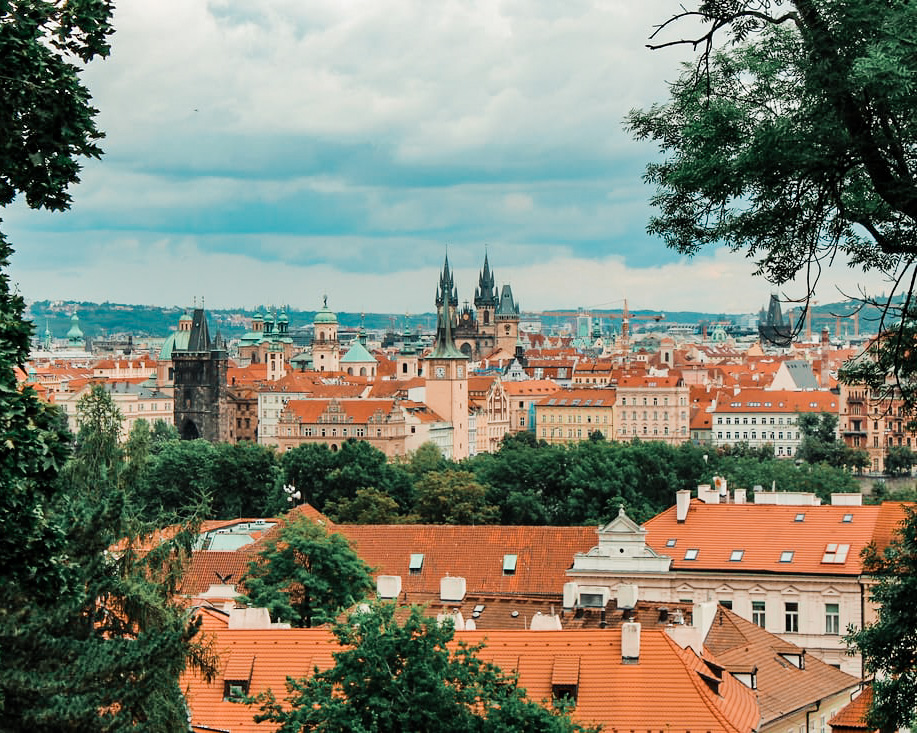 a view overlooking the city of prague with orange tiled roofs and churches in the background. many green trees all over during day time