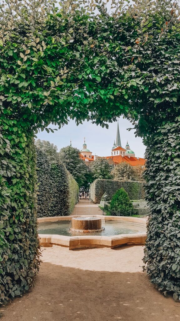 A photo of the beautiful Wallenstein Gardens with a pond and fountain surrounded by lush greenery and an archway providing a view to some of the architecture outside the gardens.