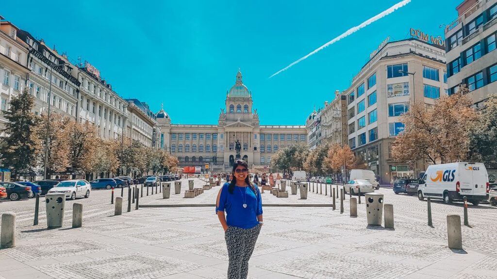 A girl standing in the center of Wenceslas Square infront of the National Museum surrounded by other historical buildings.