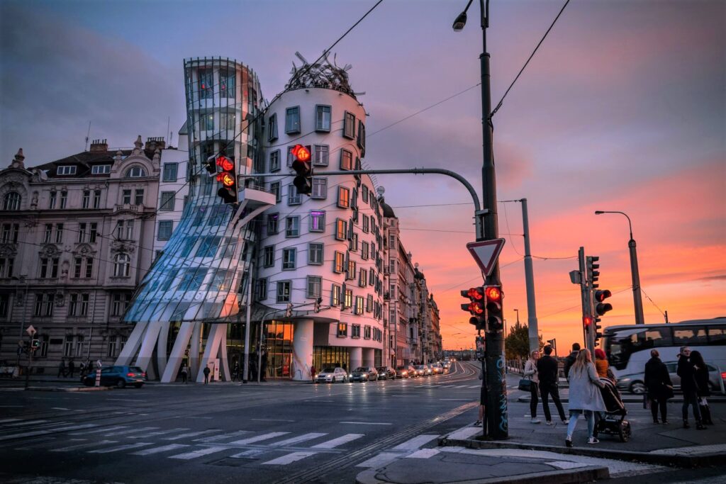 A Picture of the Dancing House at an intersection with traffic lights and cars on the road.