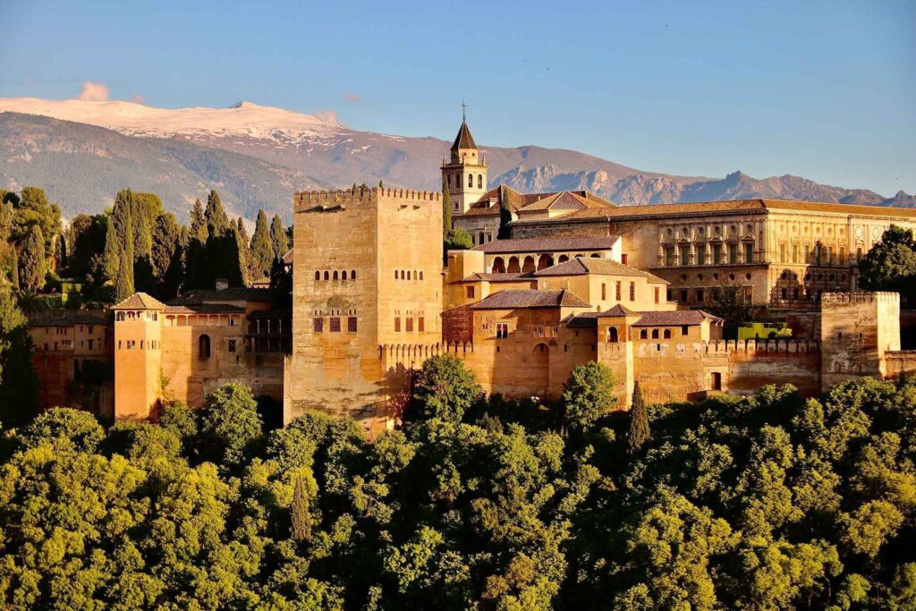 Brown concrete building complex known as the Alhambra sitting up on a height in the city of Granada with green trees surrounding the complex under blue skies. One of the most beautiful cities in Spain