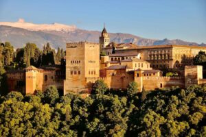 Brown concrete building complex known as the Alhambra sitting up on a height in the city of Granada with green trees surrounding the complex under blue skies