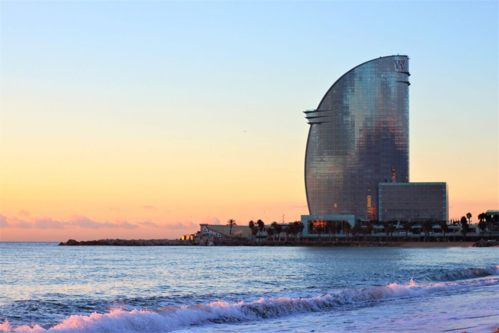 Tall curved glass building at a far corner of the beach with sand and waves nearby during sunset