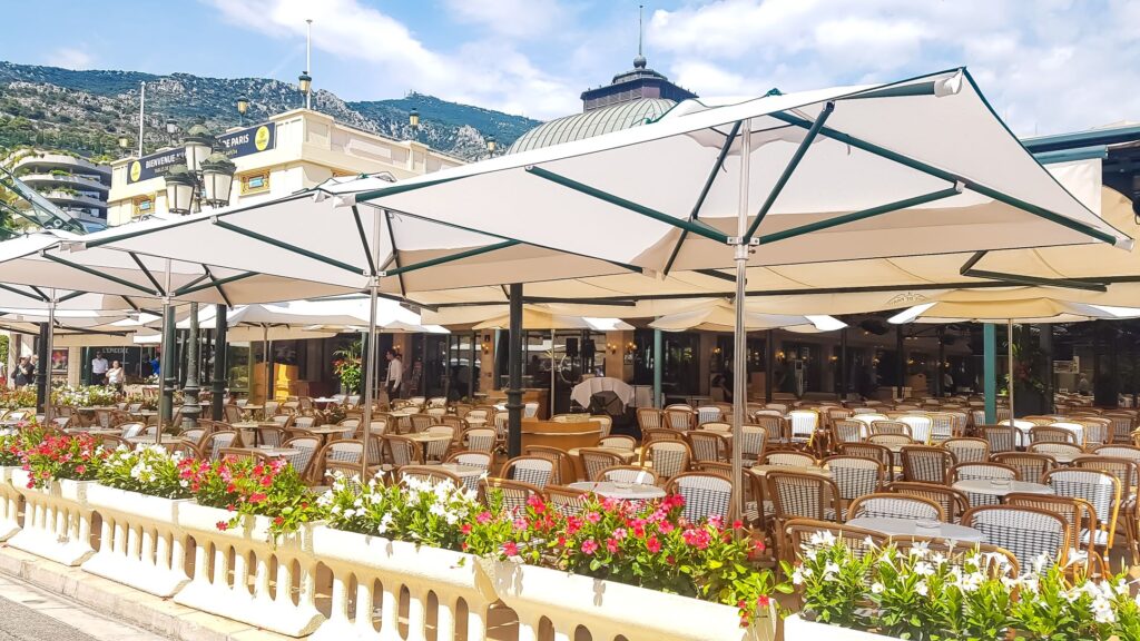 outdoor seating of the famous Cafe due Paris with tables and chairs laid out and umbrellas opened up for sun cover. The patio is line with green plants and colorful flowers