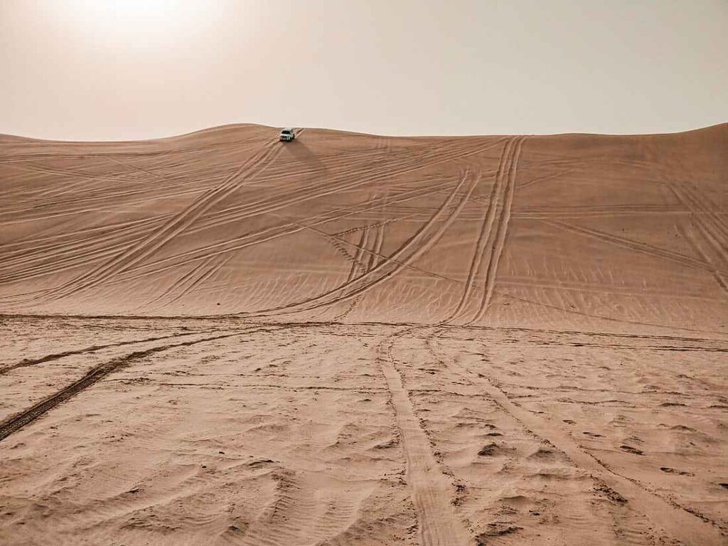 Mountains of sand with tyre tracks and a white jeep in the distance. Sun shining down on the sand