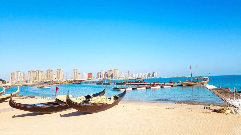 White sandy beach with wooden boats sitting on the sand as well as in the water and the skyline in a distance.A must visit place during your 3 days in Doha.