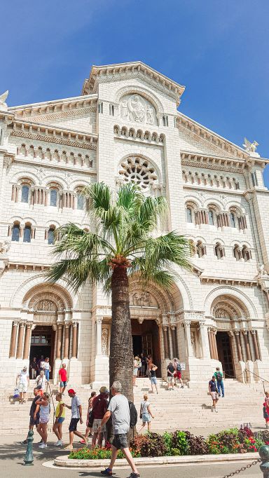 Monaco Cathedral standing tall with a palm tree in the front and plenty of people walking around the entrance