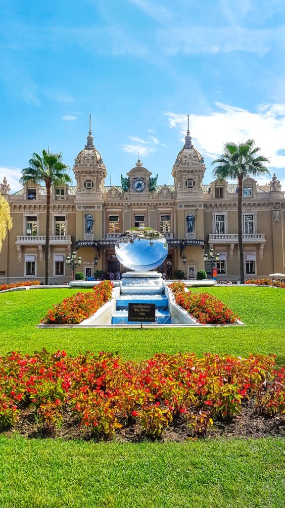 Brown concrete building known as the Monte Carlo Casino with glass globe structure infront of it sitting on a green lawn with beautifully bright flowers and palm trees lining its entrance