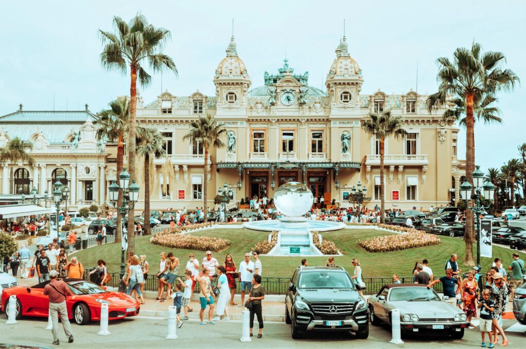 Monte carlo square with brown concrete building that is the Monte Carlo casino. A glass globe structure sitting infront of the casino on a green lawn. The square is lined with expensive cars and tourists taking lots of photos