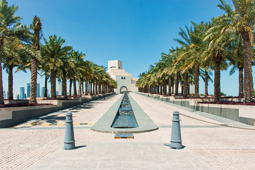 A long white concrete walkway with palm trees on either side leading to a concrete building.A must visit place during your 3 days in Doha.