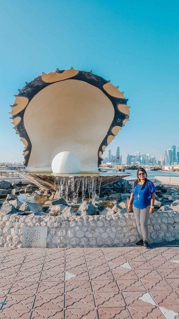 A large oyster pearl showpiece with a white pearl in the centre and fountain cascading. Girl standing in front of the monument with sea and skyline in the background