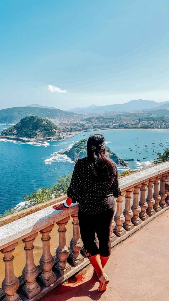 A girl standing up on top of a cliff looking out to the blue sea and some mountains covered in green trees