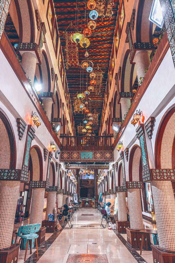 Marbled floors, Arched Walls with beautiful tiled work and ceilings with colorful lamps hanging from there. People sitting on chairs along the walkway of the interior of the Souq Waqif