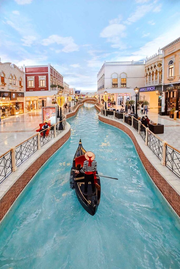 a canal inside a mall with a gondola in it and a man rowing it. shops on either side all light up and a sky painted ceiling