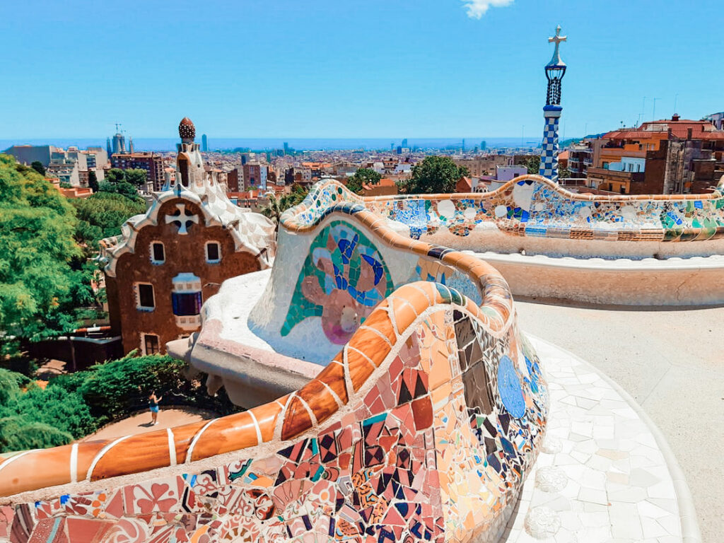 A photo from the colorful terrace at Park Güell from where you can see a gingerbread house and a view of Barcelona city in the distance.