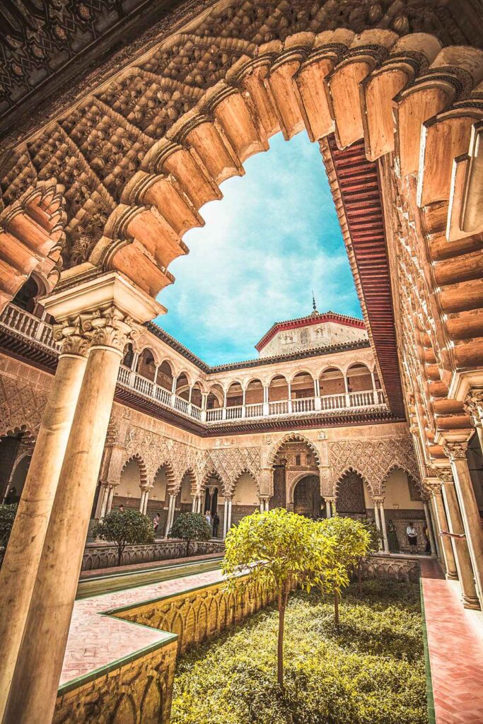 A brown carved archway with a view to a brown concrete building and a courtyard with green trees during daytime. Undoubtedly one of the top attractions in Seville