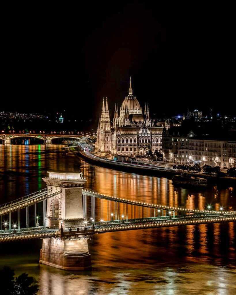 bridges over calm body of water, concrete building lit up at night. One of the best things to do in Budapest as the city lights up at night