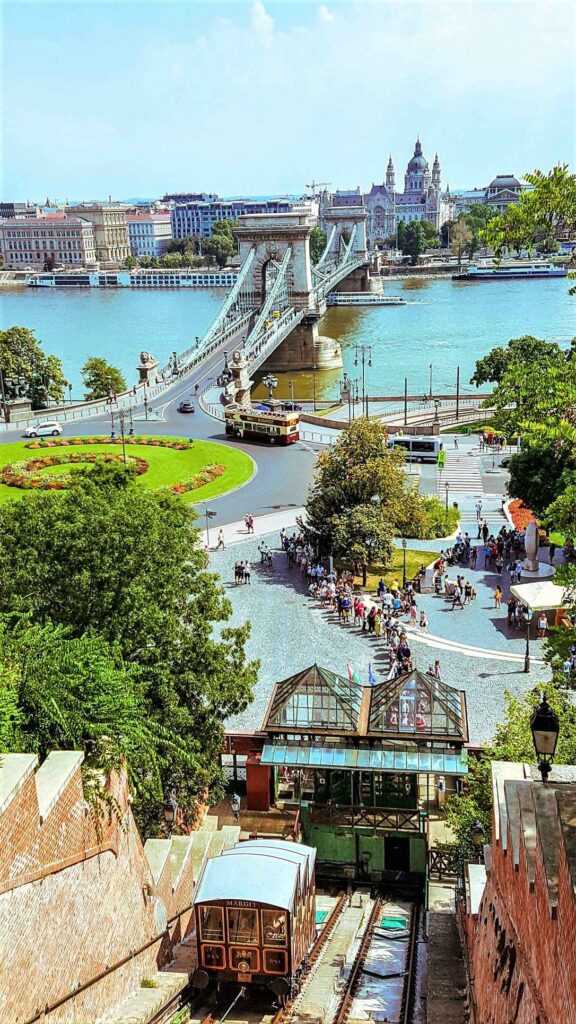 Arial view of bridge over water body, green plants all around and funicular moving down a slope. A popular activity by tourists considered one of the best things to do in Budapest