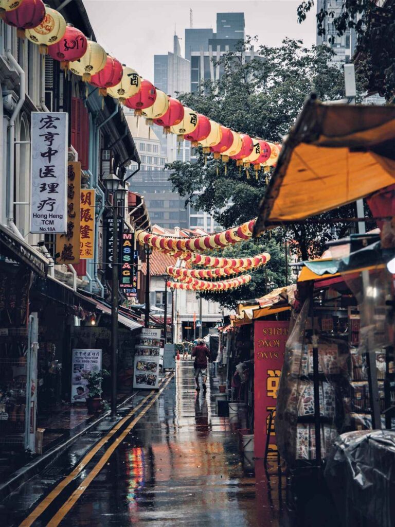 assorted red and yellow lanterns hanging above alleyway with shops of either side on a rainy day. One of the top Singapore attractions not to be missed