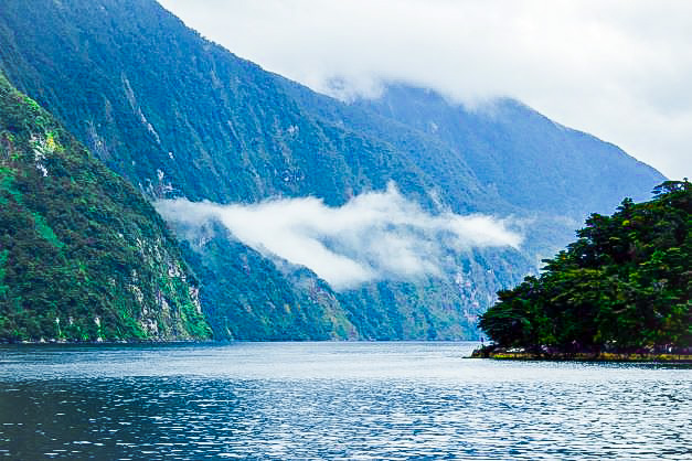 Body of water with green and brown mountains on either side and low laying white clouds