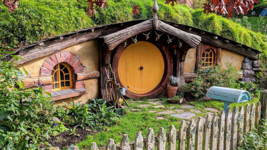 Colorful hut with wooden logs on the roof, round door and windows and wooden fence, grass in the front leading to the door and green trees. one of the top attractions in New Zealand