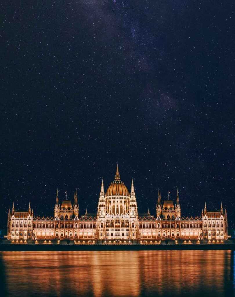 Brown concrete building lit up during night time near body of water. Viewing this stunning building is one of the best things to do in Budapest