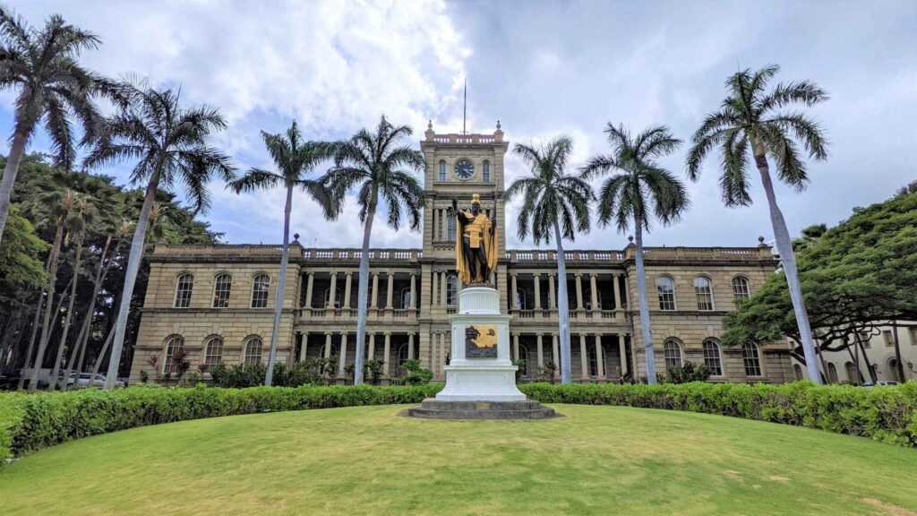 brown concrete structure sitting on green lawns surrounded by green palm treed and a famous statue infront of it all during daytime