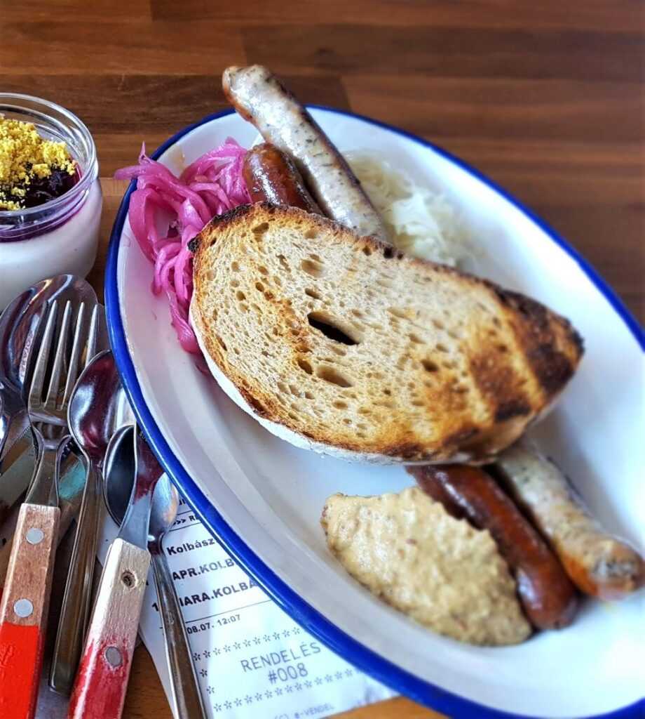 slice of bread, long saussages, pickled cabbage, in a large white dish with cutlery on the side