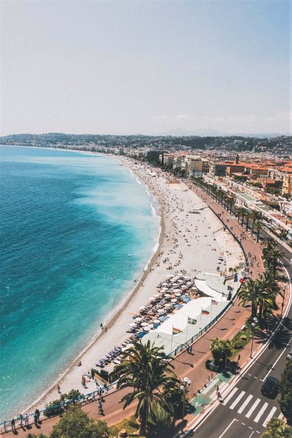 Arial view off a promenade with beach on one side, clear blue waters white sand and people sunbathing and the town centre on the other side with buildings and a concrete road. One of the top places to visit in the south of france