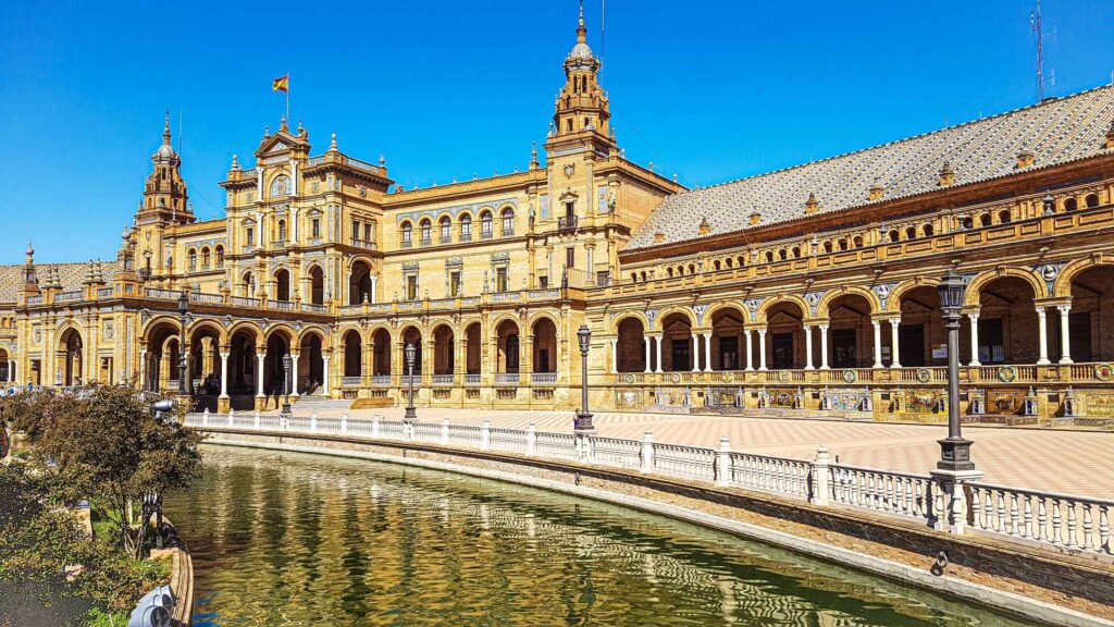 beautiful brown concrete building with long archways and tiled work and a pond infront of it. one of the top attractions in Seville