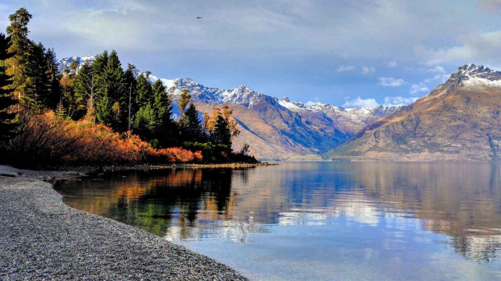 Still lake with reflection of green and brown trees, snow capped mountains and blue skies