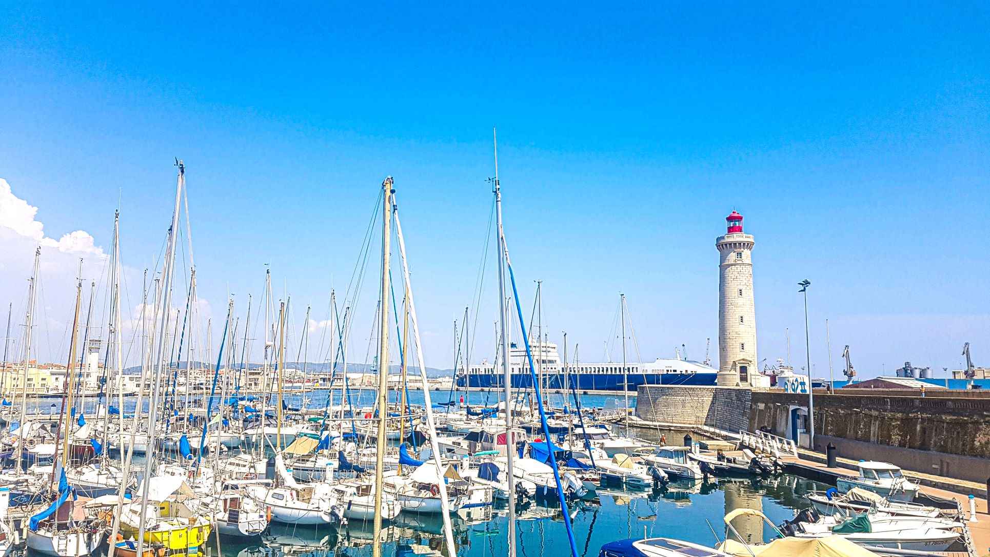 white boats with masts sitting on blue water with lighthouse in a distance