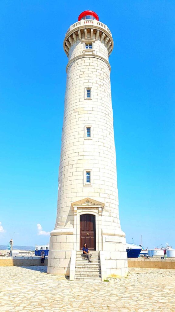 tall white lighthouse with girl sitting infront of the brown door. One of the top places to visit in the south of france