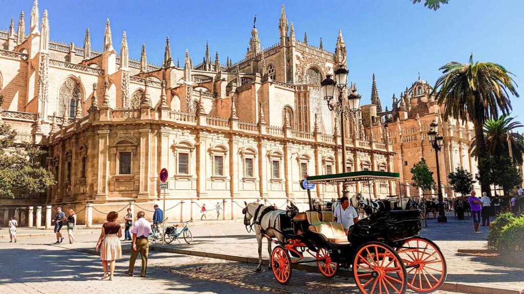 Brown concrete building with carved work and spires. Horse and carriage and people  walking on the streets that are lined with green palm trees. one of the top tourist attractions in spain.