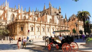 Brown concrete building with carved work and spires. Horse and carriage and people walking on the streets that are lined with green palm trees