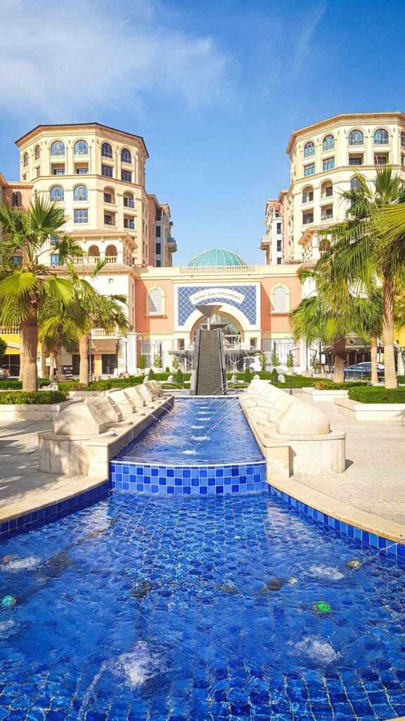 Brown concrete buildings with green trees and a blue water feature during daytime. A must visit place during your 3 days in Doha.