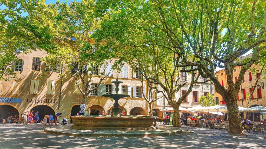 beautiful courtyard with cafes and restaurants around the perimeter with people sitting on chairs and green trees all around