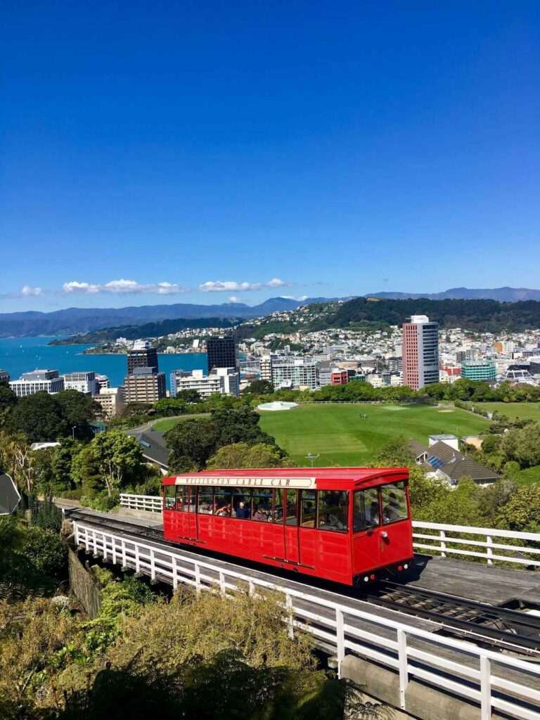 red bus moving up a concrete street with a white fence and city with buildings in the background as well as the blue sea