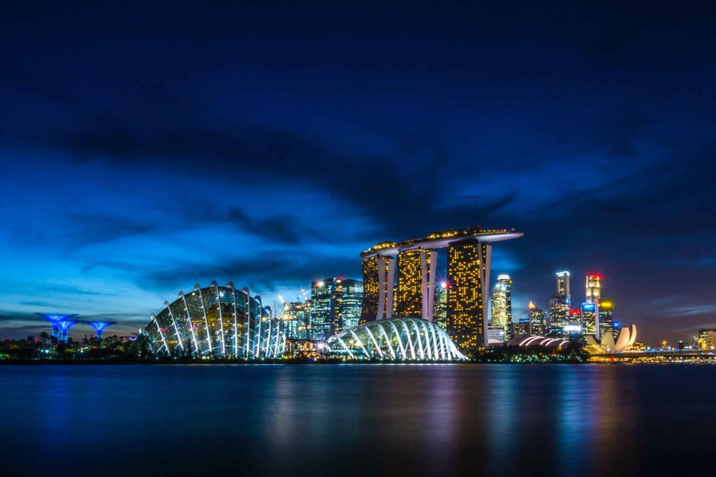 beautiful buildings lit up in a distance with calm waters in front showing a reflection of the light during night time. Marina Bay. One of the top singapore attractions not to be missed