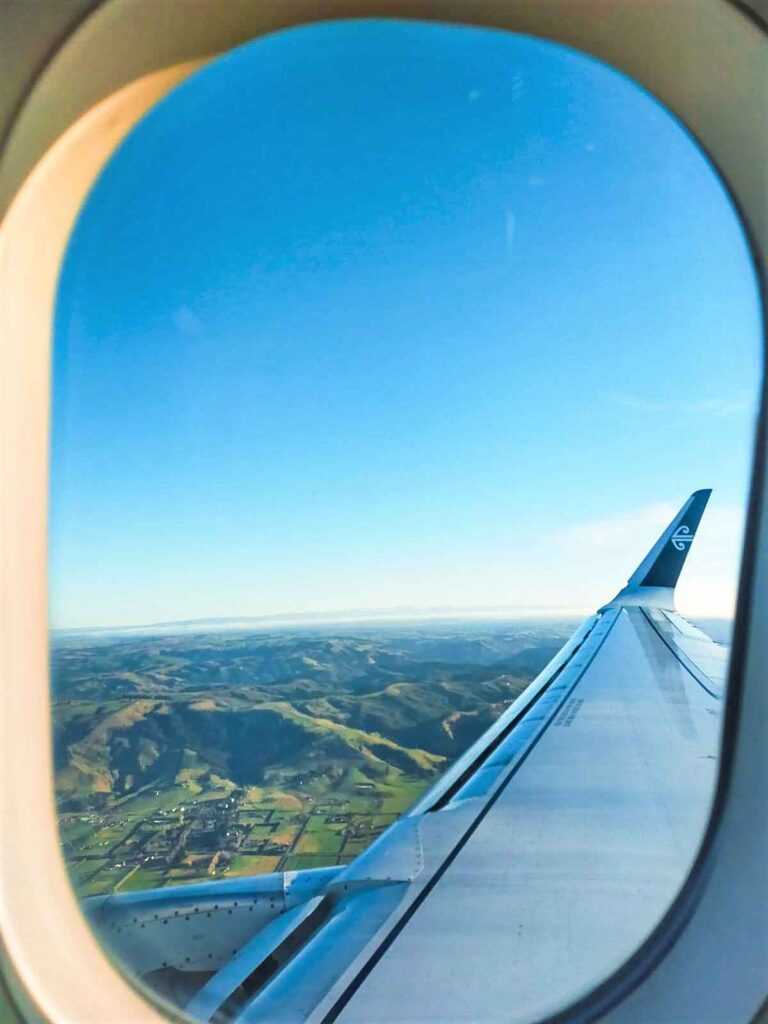 Photo of a window looking out into the sky and at the airplane wing. Booking flights in advance can save you alot of money and is one of the top travel tips